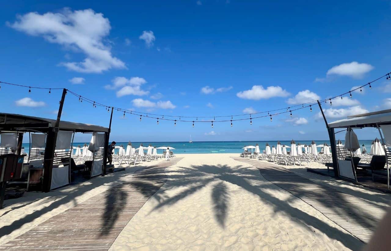 Sandy pathway lined with cabanas and string lights leads to rows of beach chairs and umbrellas facing a blue ocean under a clear sky.
